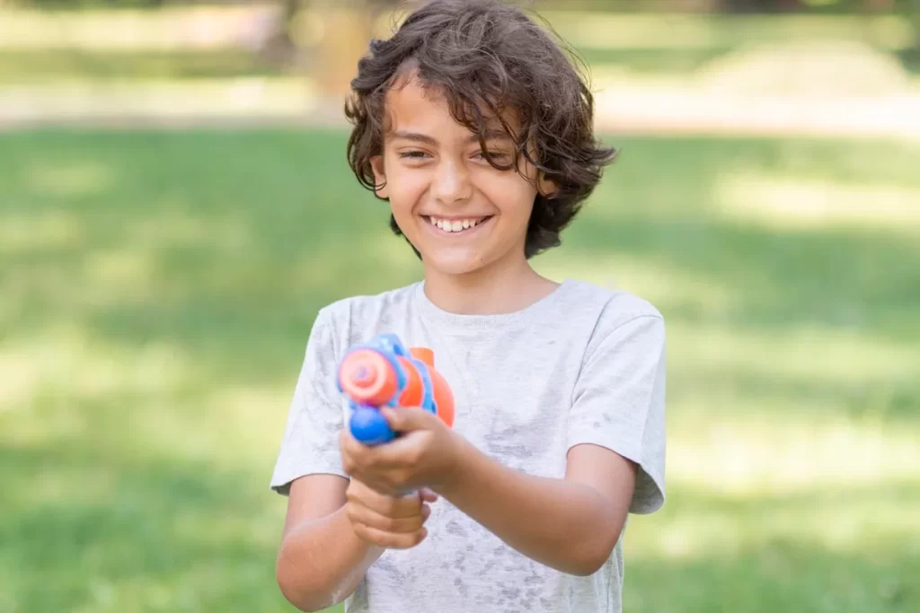 Criança sorridente com cabelo castanho e camiseta cinza brinca com pistola de água colorida ao ar livre em um parque verde.