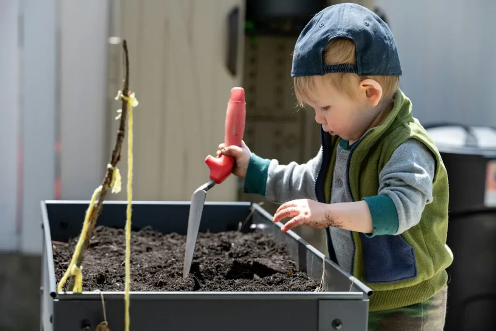 Bebê com boné azul e colete verde manuseia pá em canteiro elevado de horta em casa.