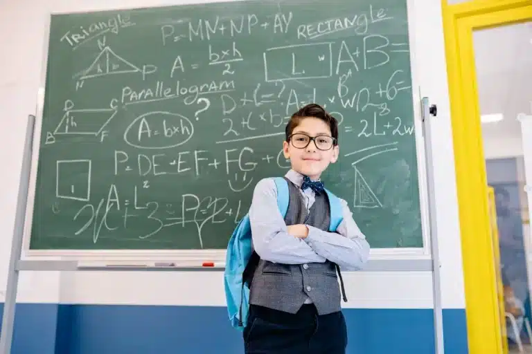 Menino sorridente de óculos e mochila em frente a um quadro-negro cheio de equações e conceitos de matemática básica.