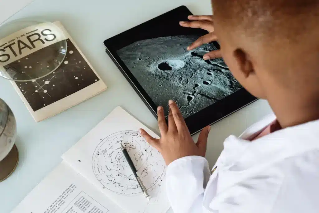Criança com jaleco usando tablet para explorar imagem da lua, livro de estrelas e mapa celeste na mesa, aprendizado interativo de astronomia.