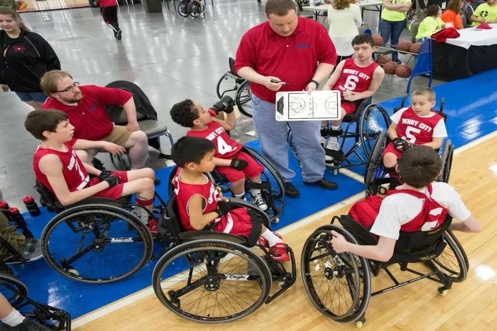 Um grupo de jovens atletas em cadeiras de rodas esportivas, vestidos com uniformes vermelhos, recebendo instruções táticas de um treinador em uma quadra de basquete adaptado, ilustrando a "mobilidade reduzida" e a prática esportiva inclusiva.