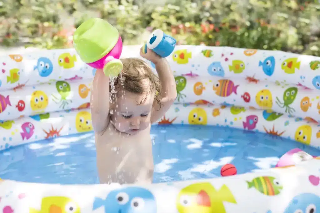 Bebê brincando com água e brinquedos em uma piscina inflável colorida decorada com peixes, ideal para o verão