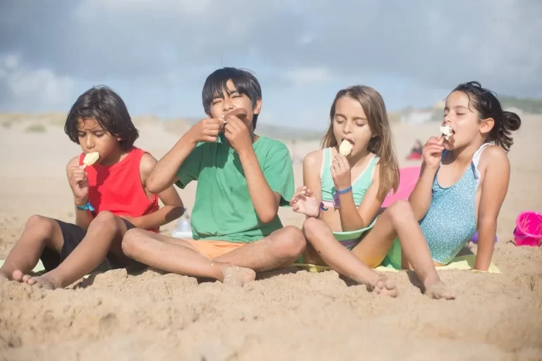 Quatro crianças multirraciais sentadas na areia da praia, aproveitando um dia ensolarado enquanto comem picolés, ilustrando um momento feliz em uma "praia para ir com criança".
