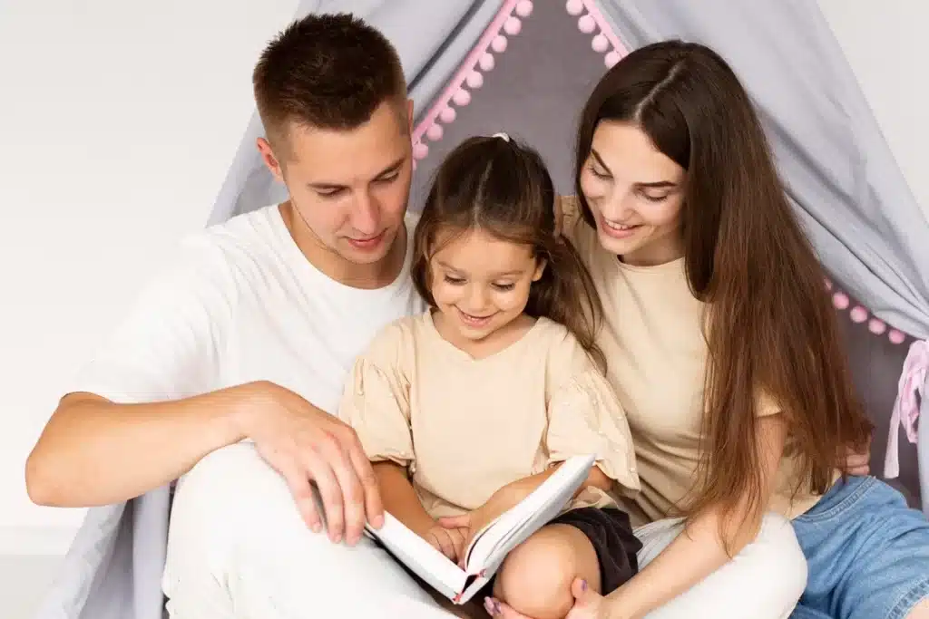 Homem e mulher sorrindo enquanto leem um livro com sua filha pequena, sentados juntos em frente a uma cabaninha infantil.