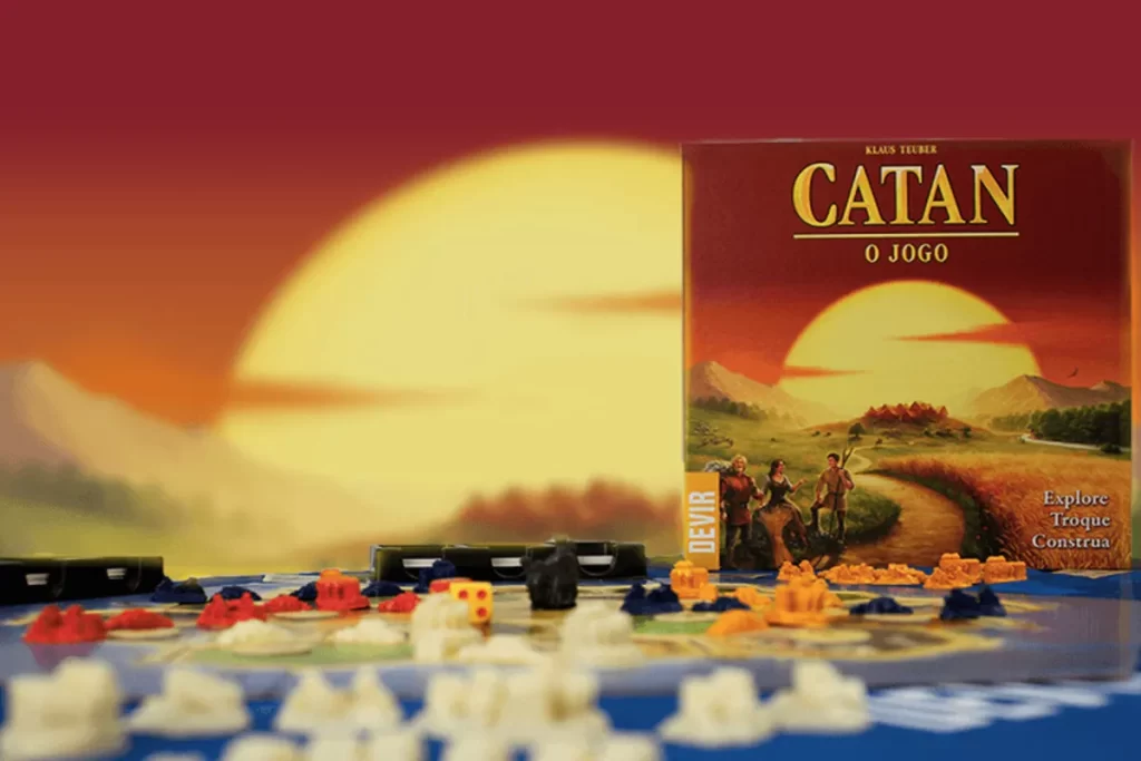 A caixa do jogo de tabuleiro Catan em primeiro plano, com o tabuleiro e as peças de jogo em segundo plano, em uma cena com um pôr do sol ao fundo.