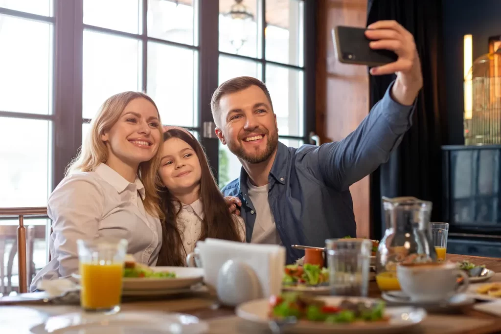 Família de três pessoas, sorrindo e tirando uma selfie em um almoço, representando a alegria e a união do momento.