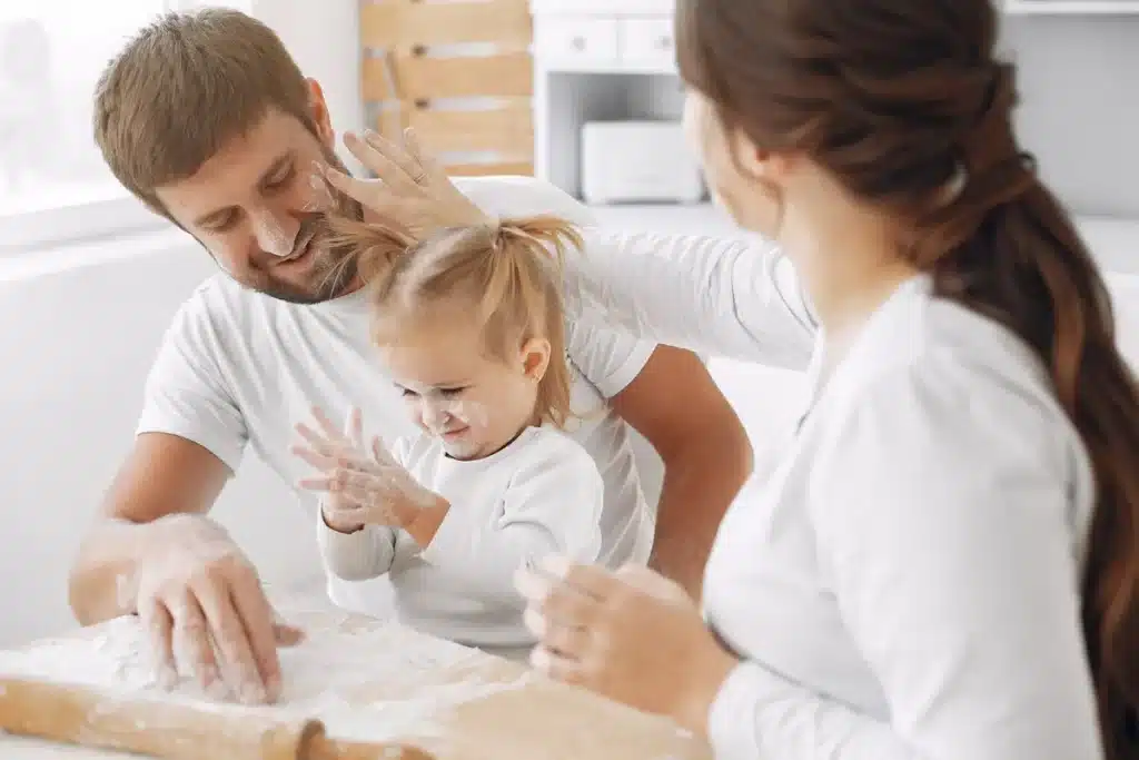 Família feliz se diverte na cozinha, em uma brincadeira com farinha enquanto preparam a receita de como fazer cookies. O pai suja o nariz da filha com farinha, e a menina ri, batendo palmas com as mãos cobertas. A imagem mostra a alegria e a união familiar na hora de cozinhar.