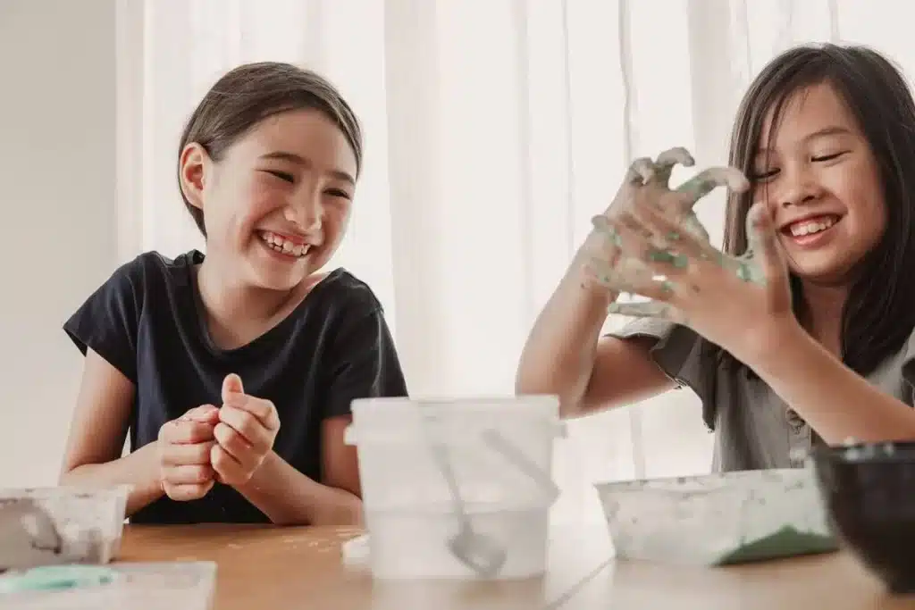 Duas crianças felizes e sorridentes brincam e misturam slime em uma mesa. A imagem ilustra o processo divertido e descontraído de como fazer slime caseiro.
