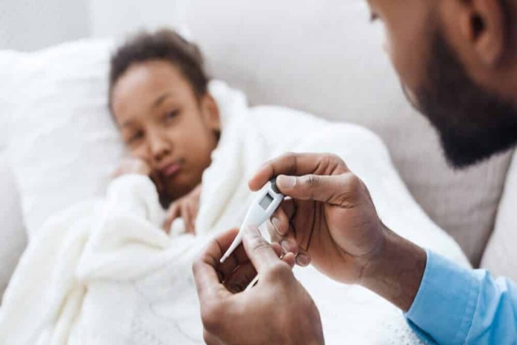 Mãos de um homem segurando um termômetro digital, com uma criança de pele escura doente e coberta por um lençol branco ao fundo.