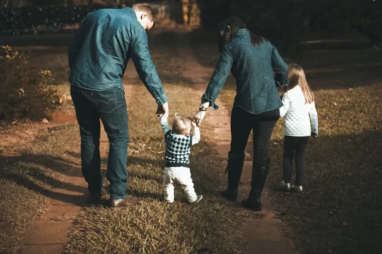 Pai e mãe, de costas, seguram as mãos de seu bebê que está dando os primeiros passos em um caminho de terra, com a filha mais velha andando ao lado.
