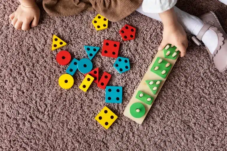 Mãos de criança brincando com jogos de encaixe de madeira coloridos no chão.