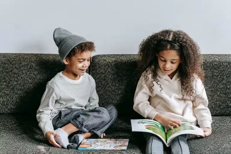 Duas crianças lendo um livro de história infantil no sofá, se divertindo com a leitura e as ilustrações do livro.