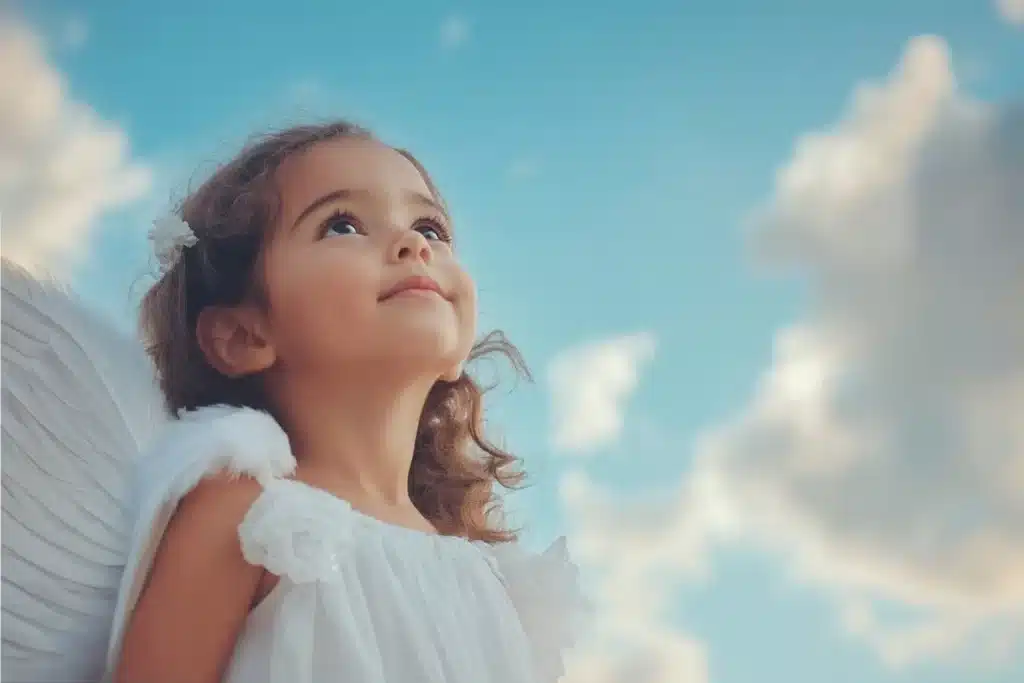 Criança pequena vestida de anjo, com asas brancas e um olhar de esperança, olhando para um céu azul com nuvens, simbolizando a inocência e a fé em Deus.