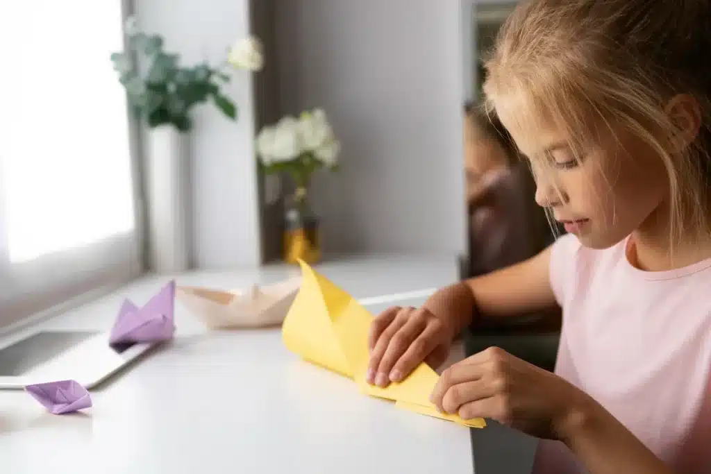 Menina loira concentrada dobrando uma folha de papel amarelo, com outras figuras de origami na mesa, simbolizando o aprendizado da arte.