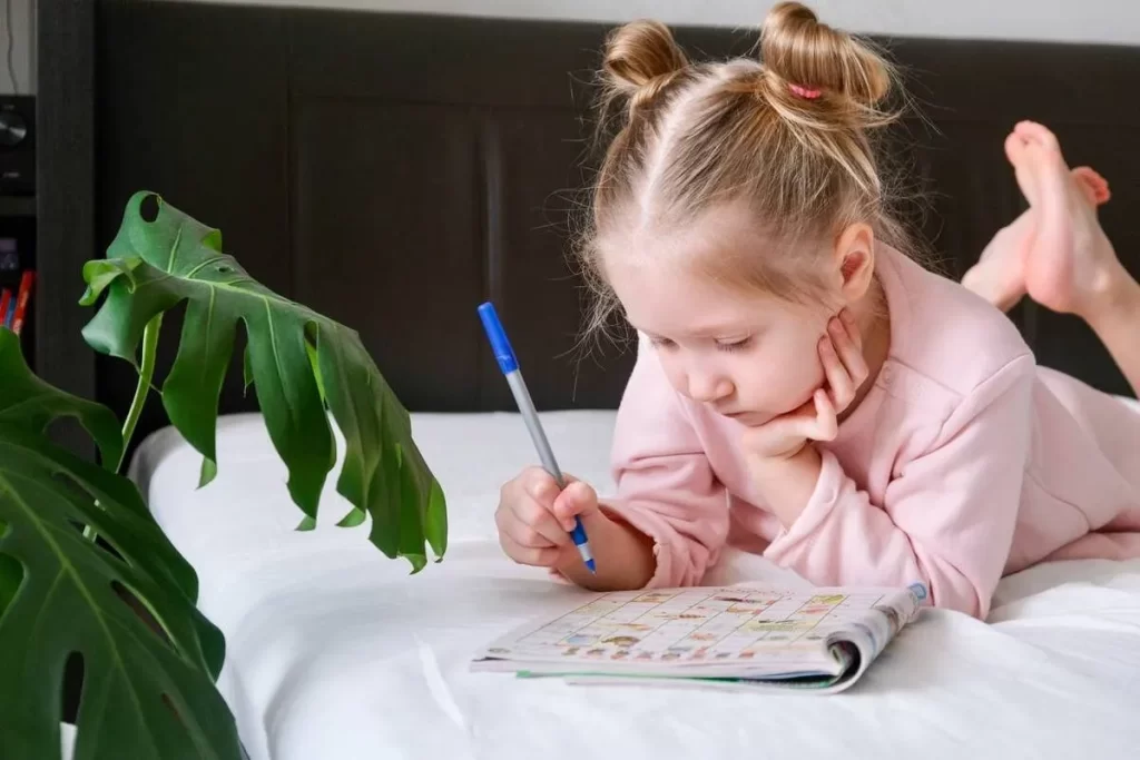 Menina sorrindo e resolvendo um jogo de palavras cruzadas deitada na cama, mostrando um momento de lazer, aprendizado e concentração.