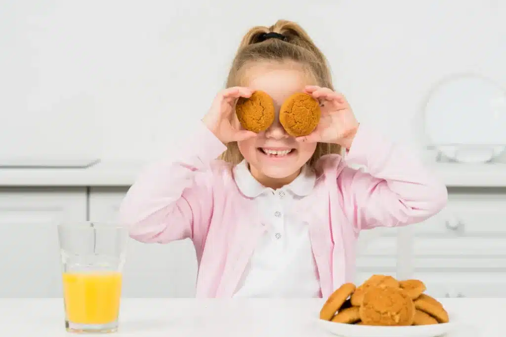 Criança se divertindo com biscoitos, colocando-os sobre os olhos, em uma mesa com um copo de suco, sugerindo um momento de lanche e alegria.