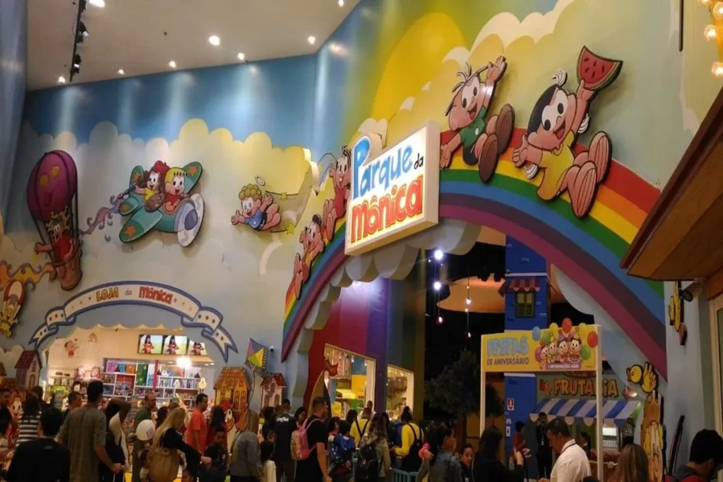 A entrada principal do Parque da Mônica, com um grande letreiro luminoso e um arco-íris colorido. As paredes são decoradas com ilustrações dos personagens da Turma da Mônica, incluindo Mônica, Cebolinha e Cascão.