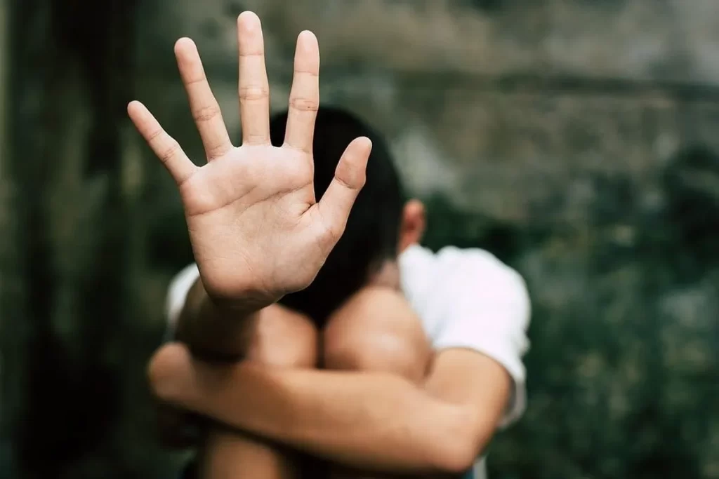 Um menino pequeno está sentado com as mãos cobrindo o rosto e os joelhos encolhidos, em uma pose que sugere medo ou vulnerabilidade. A imagem, com um fundo escuro e desfocado, representa temas de trauma e problemas psicológicos infantis (TPS).