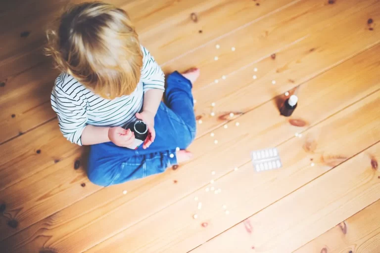 Criança sentada no chão brincando com pílulas, representando um perigoso acidente doméstico com medicamentos.