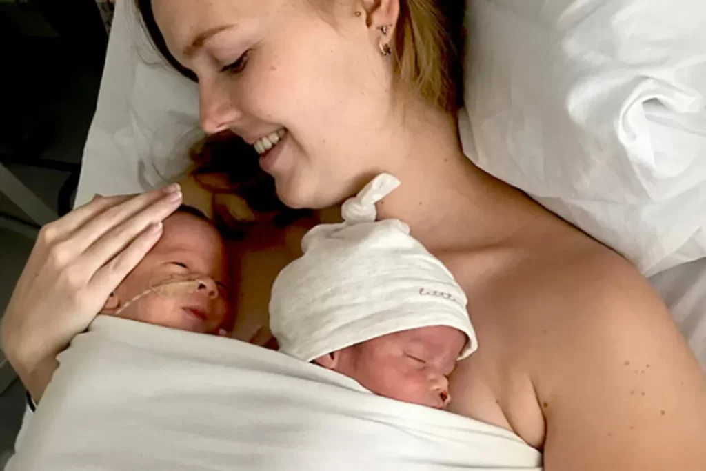 Mãe abraçando e fazendo contato pele a pele com seus gêmeos recém-nascidos em uma cama de hospital. A mãe está sorrindo, e os bebês estão enrolados em um cobertor, um dormindo com um gorro branco e o outro acordado com uma sonda nasal.