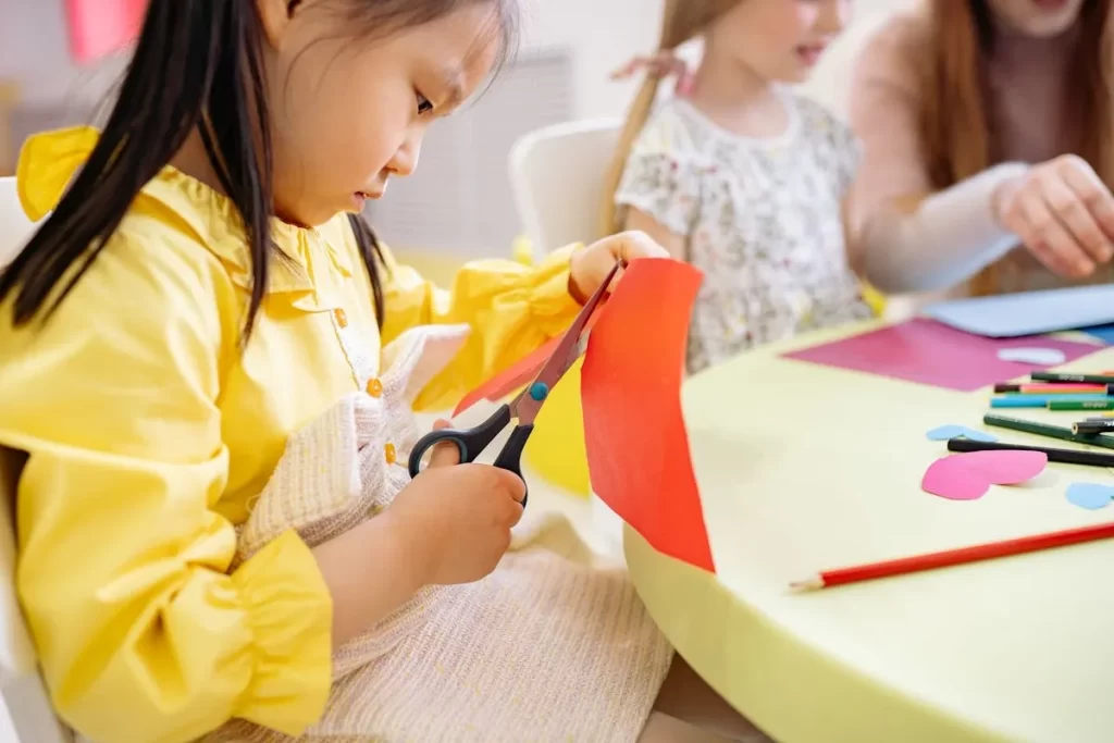 Menina concentrada cortando papel vermelho com tesoura em uma mesa de arte.