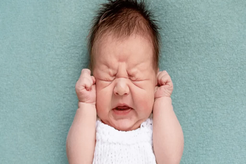 Bebê com o rosto franzido e olhos fechados, como se estivesse fazendo força, deitado em uma manta de cor turquesa, transmitindo uma sensação de desconforto.