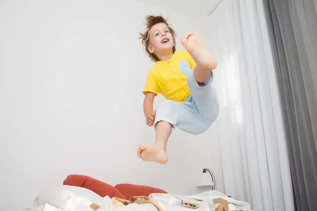 Menino de cabelos claros e sorriso alegre, vestindo camiseta amarela e calça clara, pulando no ar sobre uma cama em um quarto bem iluminado, em uma representação visual da energia característica da criança hiperativa ou simplesmente de uma criança brincando e se divertindo.