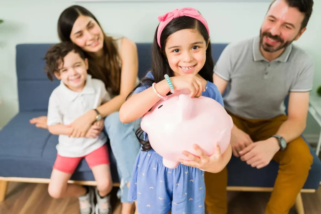 Menina sorridente segurando um cofrinho rosa, com a família sorrindo ao fundo, em uma lição de economia.