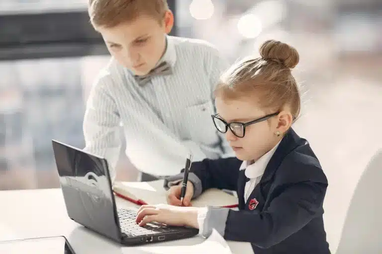 Crianças vestidas de forma formal usando laptop e caderno, representando economia doméstica, planejamento financeiro e educação infantil.