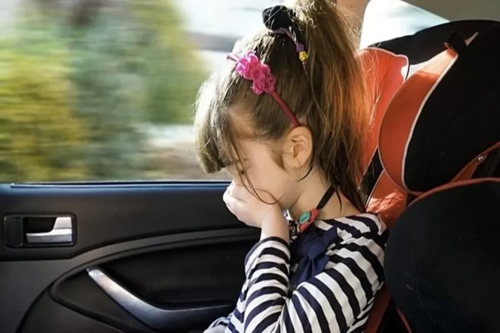 Criança no banco de trás do carro com sinais de enjoo de movimento, com a cabeça baixa e os olhos fechados.