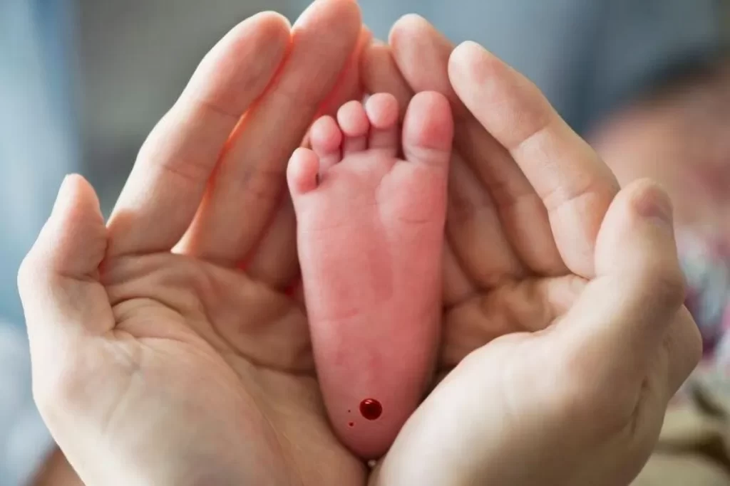 Duas mãos de adulto seguram o pé de um recém-nascido com uma gota de sangue no calcanhar, representando o exame do pezinho.