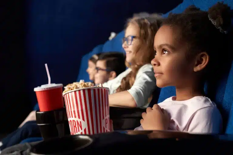 Crianças assistindo filme no cinema com pipoca e refrigerante.