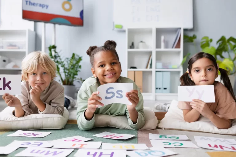 Três crianças sentadas em almofadas no chão, segurando cartões com letras do alfabeto durante uma aula de inglês.