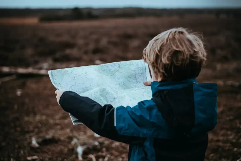 Uma criança de costas segurando um mapa desdobrado, aparentemente perdida ou planejando um caminho. A imagem mostra a criança no meio de um campo de terra, em um dia nublado.