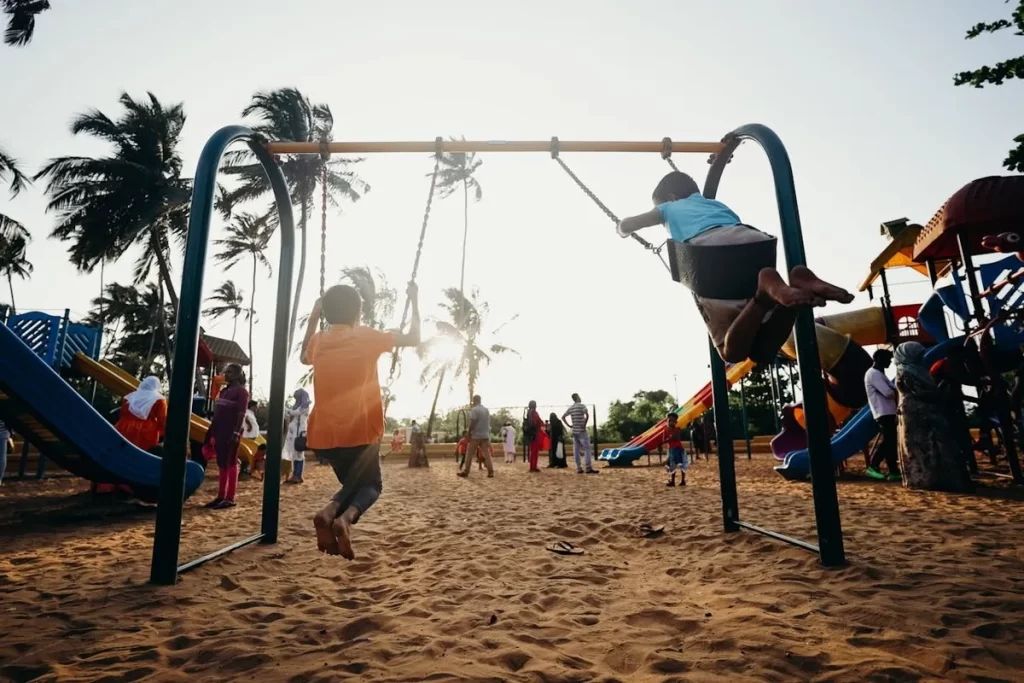 Dois meninos brincando de balanço em um playground de areia ao pôr do sol.