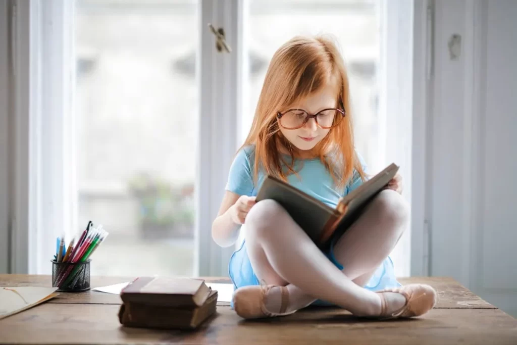 Menina com cabelos ruivos usando óculos e lendo um livro em uma mesa de madeira.
