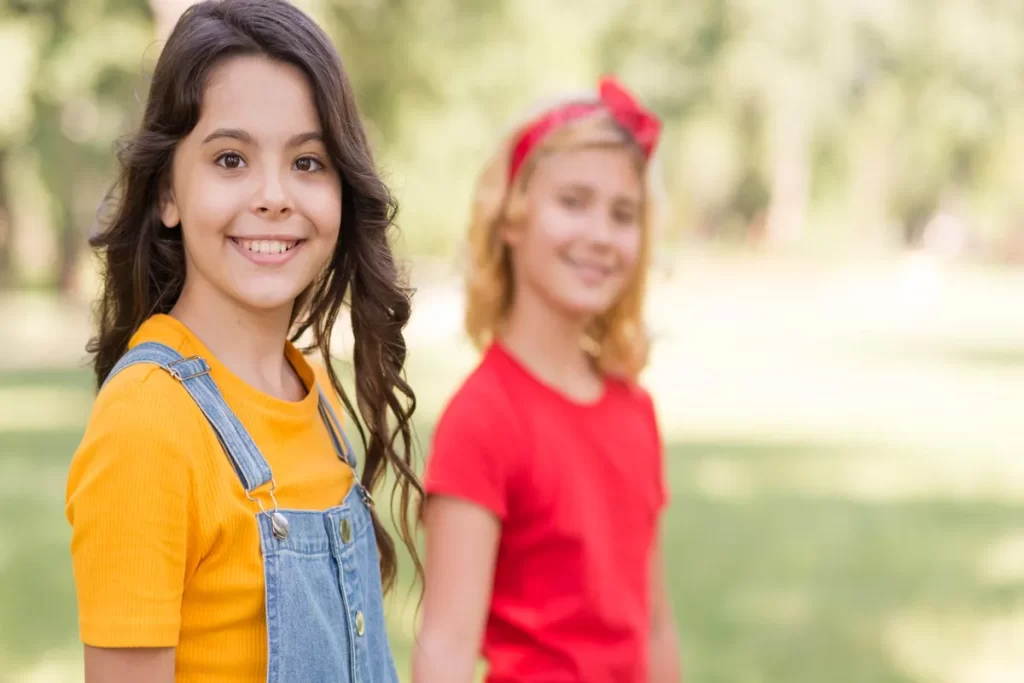 Duas meninas pré-adolescentes em um parque. A menina em primeiro plano, em foco, veste uma camiseta amarela e uma jardineira jeans. A outra, desfocada ao fundo, usa uma camiseta vermelha e uma faixa de cabelo vermelha, com um sorriso.