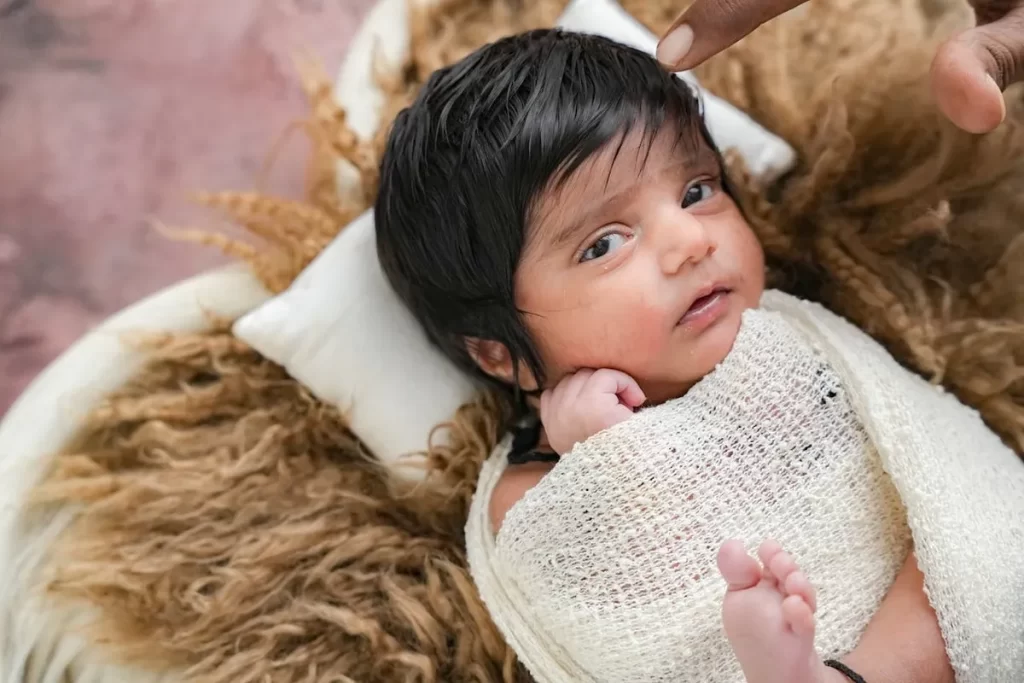 Um close-up de um bebê recém-nascido, com cerca de dois meses, deitado de costas. O bebê, que tem cabelo escuro e está parcialmente coberto por um pano branco, olha diretamente para a câmera. O cenário inclui uma manta de pele e um dedo adulto tocando suavemente a cabeça do bebê. A imagem foca na expressão curiosa e no rosto do bebê.