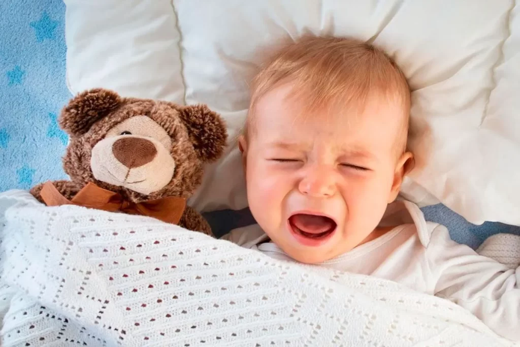 Um bebê chorando intensamente deitado na cama, com a boca aberta e os olhos fechados. Ele está coberto com uma manta branca, e um urso de pelúcia marrom está ao seu lado. A imagem simboliza o choro noturno e o desconforto que o terror noturno pode causar.