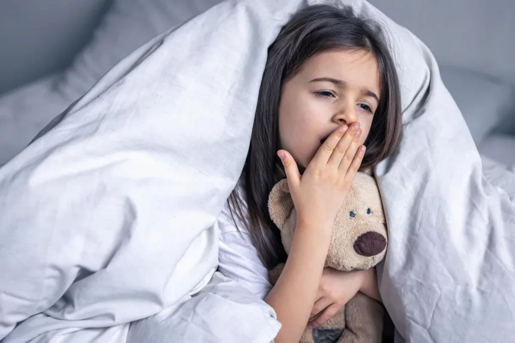 Uma menina bocejando em sua cama, com uma expressão sonolenta, enquanto se esconde sob um edredom branco e abraça um urso de pelúcia. A imagem ilustra o cansaço e a fadiga associados a transtornos do sono infantil.