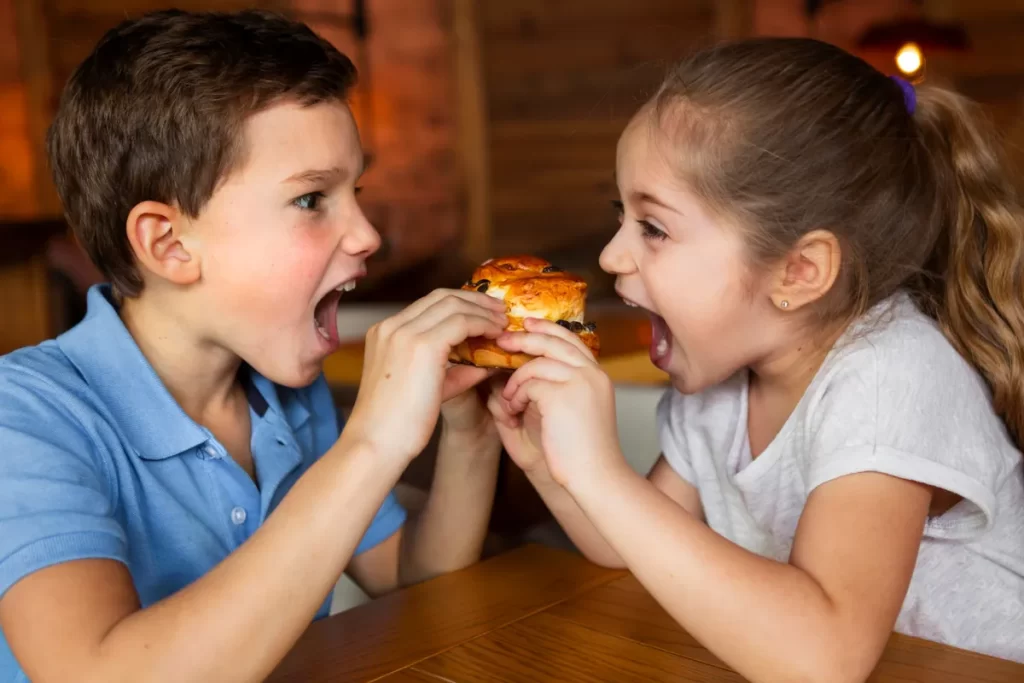 Dois irmãos, um menino e uma menina, em uma mesa de madeira, abocanhando simultaneamente um pequeno pão ou hambúrguer em miniatura. Ambos estão sorrindo com a boca aberta, olhando um para o outro, em um momento de diversão e competição por um alimento, provavelmente um petisco ultraprocessado ou fast food. O menino usa uma camisa polo azul e a menina, uma camiseta branca.