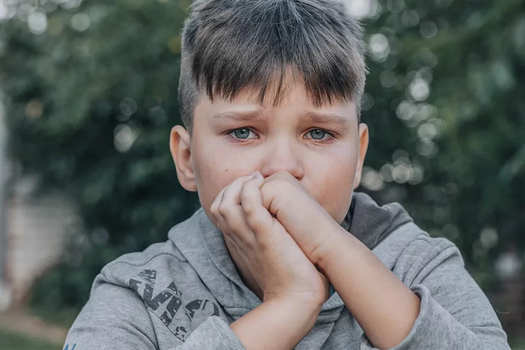 Menino pré-adolescente com olhos azuis cobrindo o rosto com as mãos, expressando tristeza ou ansiedade.