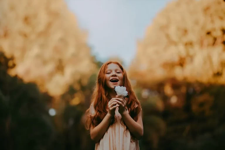 Retrato de uma menina de cabelo ruivo, longo e ondulado, com um sorriso largo e olhos brilhantes, exalando autoconfiança e felicidade. Ela está em um parque ou floresta, segurando uma flor branca perto do rosto. O plano de fundo (bokeh) mostra árvores com folhagem outonal amarelada, criando um cenário dramático e suave com iluminação contrastante.