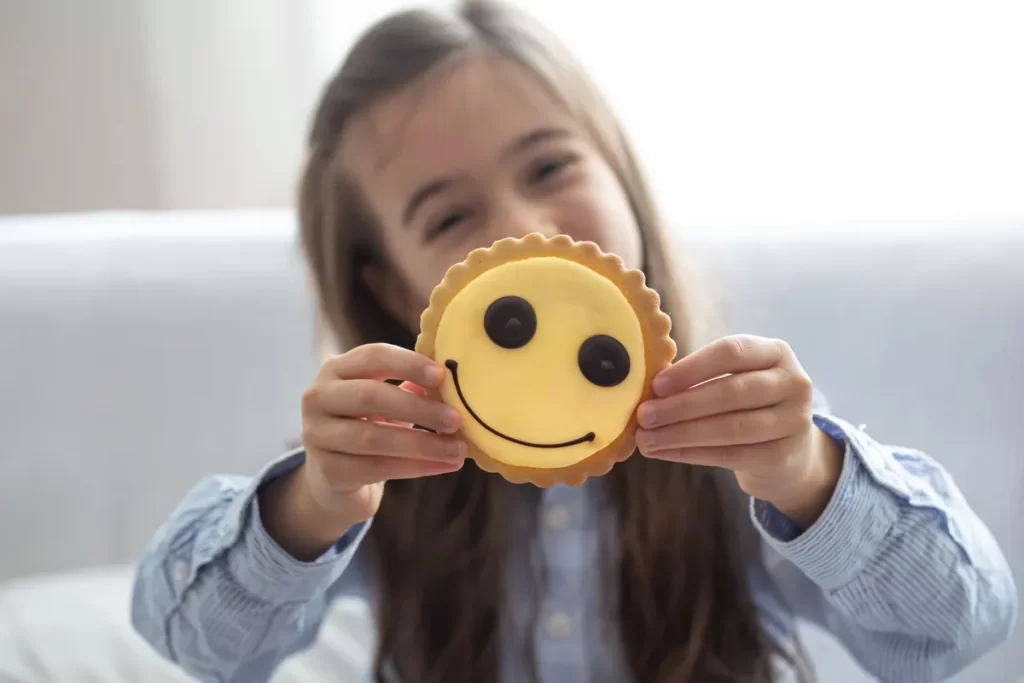 Close-up de uma menina sorridente, com cabelo castanho comprido e camisa azul listrada, segurando um grande biscoito redondo com cobertura amarela e um rosto feliz desenhado em chocolate. O biscoito, que tem uma borda de massa frisada, está no centro do foco, simbolizando a recompensa, a espera ou o autocontrole em relação a guloseimas.