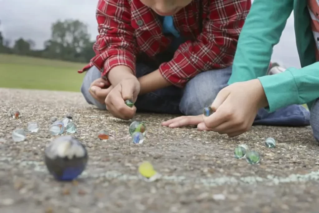Duas crianças ajoelhadas no chão jogando bolinha de gude, focadas nas bolinhas de vidro coloridas no asfalto.