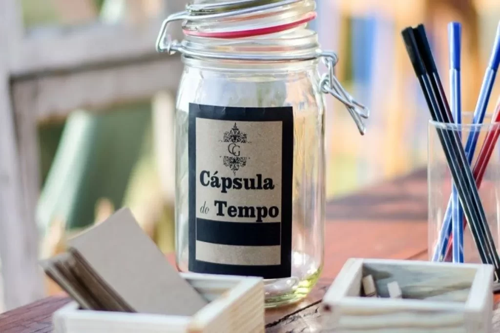 Um pote de vidro transparente com tampa de metal e vedação vermelha, rotulado com uma etiqueta rústica em preto e kraft escrito "Cápsula do Tempo". O pote está sobre uma mesa de madeira rústica, cercado por pequenos cartões de papel kraft em uma caixa de madeira e um copo de vidro com canetas e lápis coloridos, sugerindo um projeto de memória e mensagens.