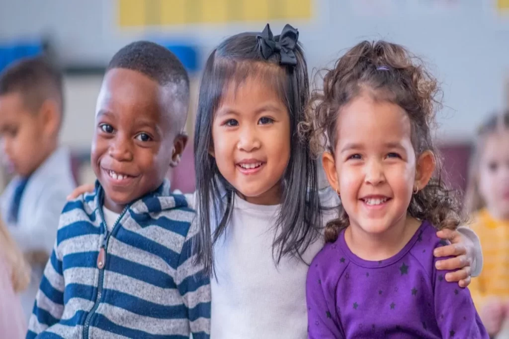Três crianças de etnias diferentes sorrindo e abraçadas, representando amizade e diversidade em um ambiente escolar.