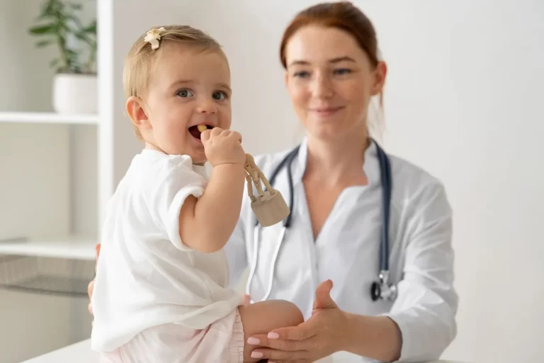 Médica pediatra sorrindo examinando um bebê feliz que está mordendo um brinquedo de madeira durante a consulta.
