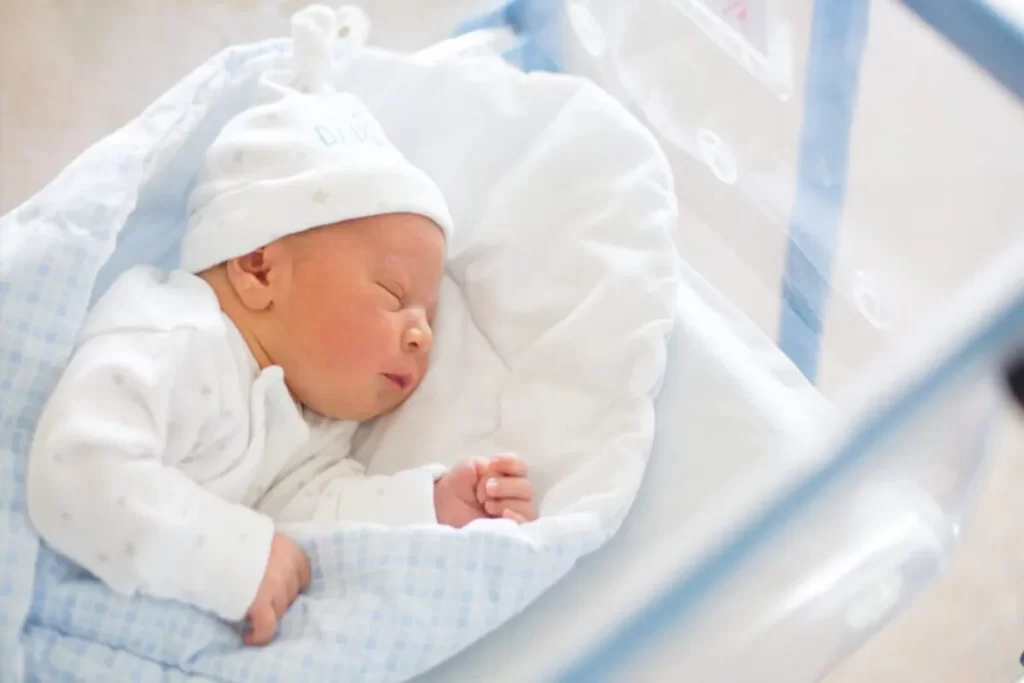 Close-up de um bebê recém-nascido dormindo em um berço hospitalar (bassinet), enrolado em um cobertor azul e branco e usando uma touca branca, em um ambiente hospitalar.