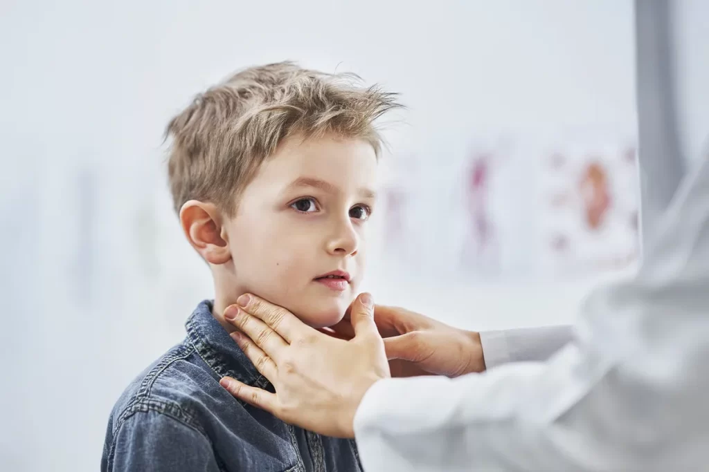 Close-up lateral de um médico ou pediatra (vestindo um jaleco branco) examinando o pescoço de um menino (criança, 5-7 anos, de cabelo loiro e vestindo uma camisa jeans). As mãos do profissional tocam delicadamente a área do pescoço, indicando a palpação das glândulas e linfonodos, procedimento comum para diagnosticar condições como faringite ou amigdalite.